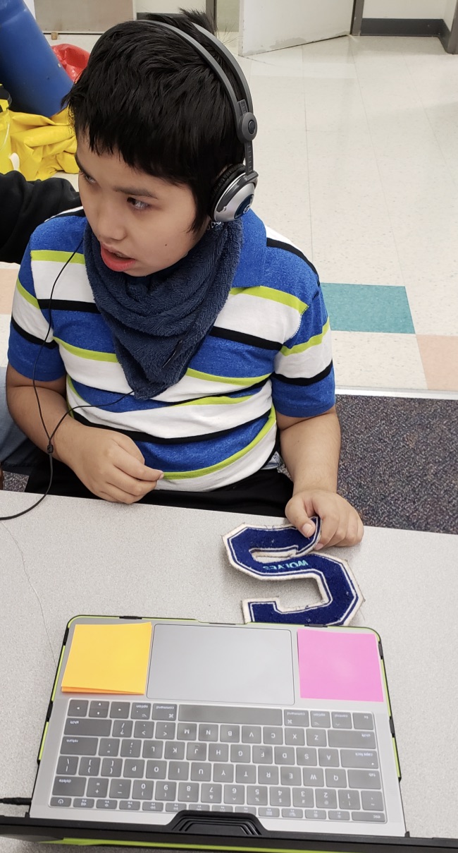 A young boy wearing headphones is listening to a book on the computer as he looks into the hallway.