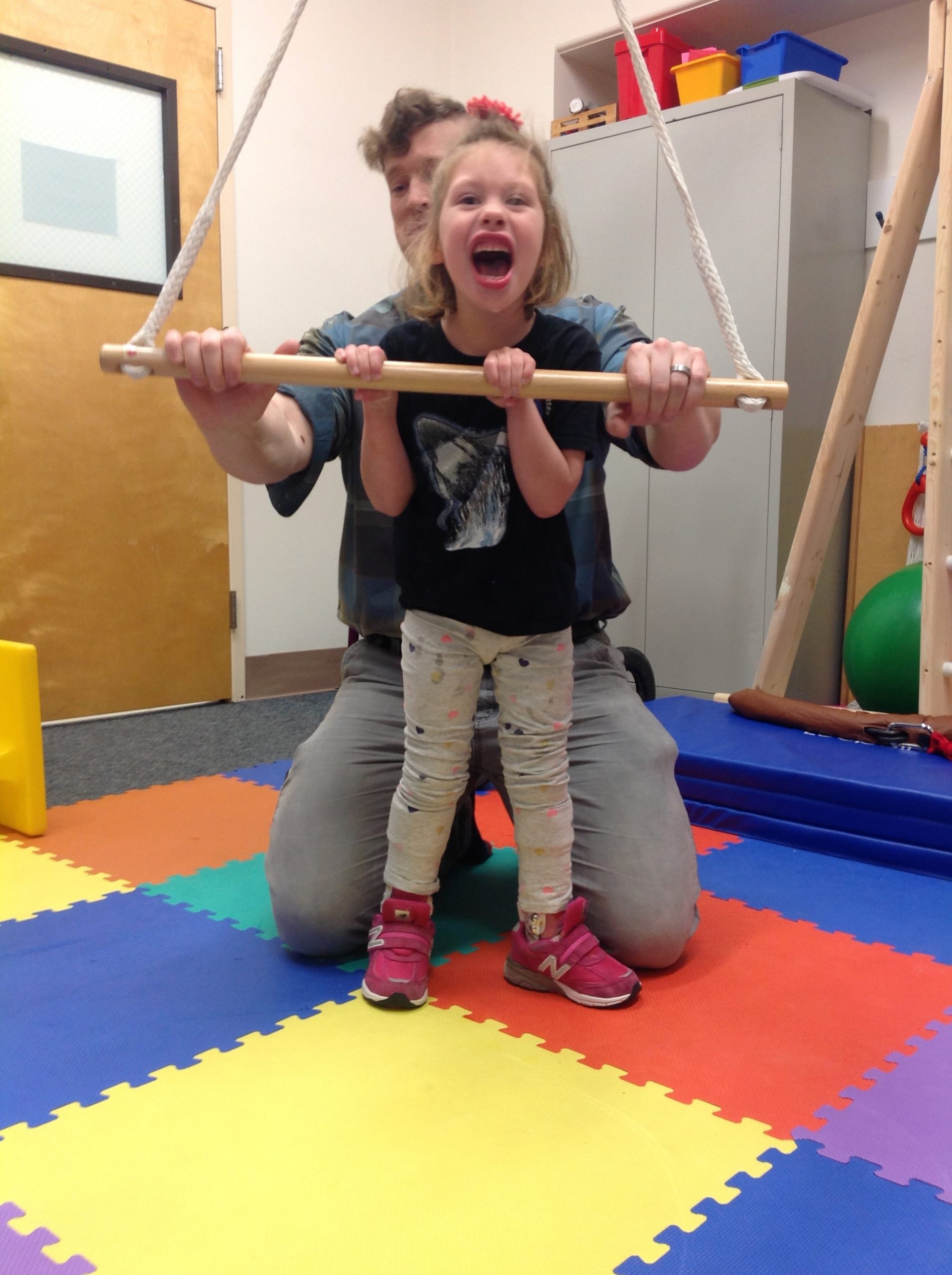 A young girl is using bar supports to stand tall while her teacher kneels behind her.
