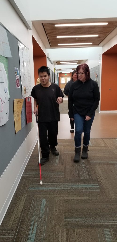 Young man with dark hair, wearing dark clothes is walking down a hall with a white mobility cane beside a woman with red hair, wearing dark clothes.